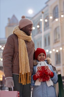 Mutlu siyah baba ve kızının dikey portresi Noel alışverişinin keyfini çıkarıyor kışın açık havada hediyelerle yürüyor.
