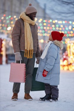 Mutlu siyah baba ve çocuğun dikey portresi Noel alışverişinin keyfini çıkarıyor ve kışın açık havada birbirlerine bakıyorlar.