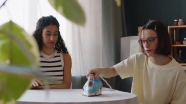 Beyinsel felçli beyaz bir kızın, çift ırklı bir bayan arkadaşıyla sohbet ederken, mutfak masasındaki masa örtüsünü sıcacık bir dairede ütülerken orta boy fotoğrafı.