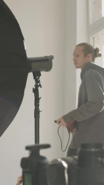 Genç beyaz erkek fotoğrafçı asistanının, stüdyo içinde fotoğraf çektirmek için ışıklandırma ekipmanı hazırlarken çekilmiş dikey mercek görüntüsü.