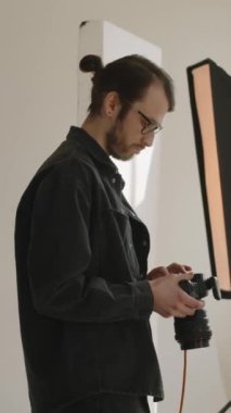 Dikey yan görüntü, genç beyaz erkek fotoğrafçı elinde dslr kamerayla duruyor ve profesyonel fotoğraf stüdyosunda çalışırken görüntülere bakıyor.
