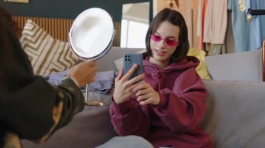 Serebral felçli, pembe y2k gözlük takan ve Melez kız arkadaşı elinde ayna tutarken ve evde birlikte eğlenirken akıllı telefondan komik bir selfie portresi çeken beyaz bir kız.