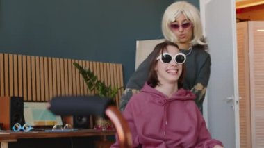 Çift ırklı, peruklu, beyaz, pembe gözlüklü arkadaşını sandalyeye oturtup gülen orta yaşlı bir genç kız. Evde eğlenen iki kız. Kızlardan biri beyin felci geçiriyor.