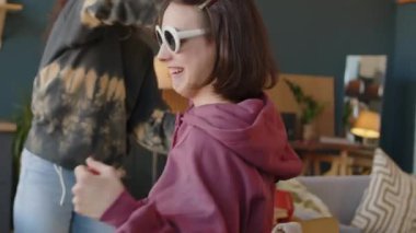 İki komik çoklu etnik kökenli genç kızın şık kıyafetler giyip modern dairede sandalyede dönerek eğlendiği orta boy bir fotoğraf. Kızlardan biri beyin felci geçiriyor.