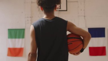 Kolunun altında topu tutan çift taraflı erkek basketbolcunun arka plan yavaşlaması. Beyaz duvarda Birleşik Krallık bayrağı asılı iç saha boyunca sepete doğru yürüyor.