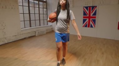 Kendine güvenen, basketbol şortlu, kolunun altında basketbol oynayan kapalı sahada kameraya poz veren iki ırklı kızın yavaş yavaş portresi.