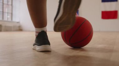 Basketbolun alçak kesimi parke zemininde seçici bir odak noktasındayken iki yüzlü genç basketbolcu ellerinde toplarla iç saha antrenmanı için sahaya geliyorlar.