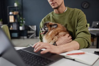 Kucağında sevimli bir köpek tutan ve evcil hayvan dostu fotokopi odasında çalışırken bilgisayar kullanan tanınmamış bir adamın yakın çekimi.
