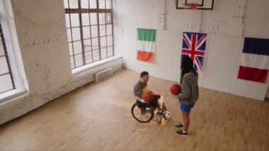 Tekerlekli sandalyede oturan, basketbol topuyla serbest atış talimi yapan, çift ırklı bayan antrenörle iç saha eğitimi alan, engelli beyaz bir adamın geniş açılı çekimlerinden.