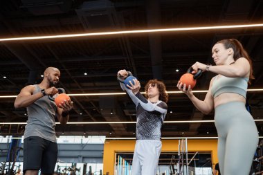 Spor salonunda ağırlık kaldırma eğitiminde müşterilere yardımcı olan kaslı siyahi bir adamın düşük açılı portresi.