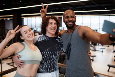 Spor salonunun içinde fotoğraf çektirirken kameraya gülümseyen ve birlikte poz veren üç genç insanın belden yukarı portresi