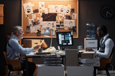 İki polis dedektifinin yan görünüm portresi. Ofiste karşılıklı taraflar halinde oturmuş dava araştırması yapıyorlar.