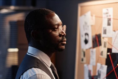 Siyahi erkek dedektifin olayı araştırırken kanıt panosuna baktığı dramatik bir yan bakış açısı.