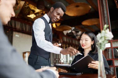 Siyahi genç adamın portresi, kadın fırtınasından emir alan ve lüks restorandaki menüye işaret eden, boşluğu kopyalayan bir garson.