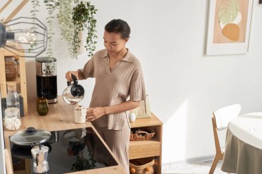 Side view portrait of young Black woman making coffee at home in morning in simple kitchen interior, copy space clipart