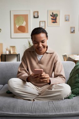 Sosyal medyada rahat koltukta bacak bacak üstüne atarak otururken akıllı telefon kullanan gülümseyen zenci kızın tam boy portresi.