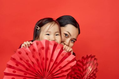 Stüdyoda kırmızı arka planda kağıt yelpaze tutan Asyalı bir annenin portresi.