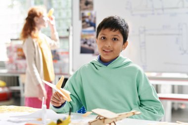 Portrait of smiling boy in hoodie gluing airplane model at desk in aeromodeling class clipart