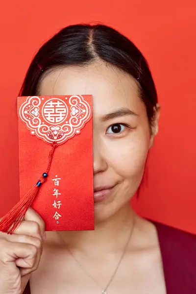 stock image Vertical half portrait of young Asian woman holding hongbao envelope against red background and looking at camera Translation Have overflowing abundance hundred years