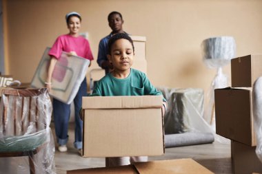 Sevimli küçük çocuk paketlenmiş karton kutu taşıyor ve ailesine taşınmalarında yardım ederken diğerinin üstüne koyuyor.