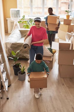 Sevimli küçük çocuk, ailesinin önünde rulo halı ve paket destesiyle taşınırken yanında ev malzemeleriyle dolu bir kutu taşıyor.