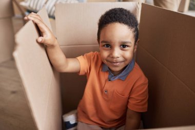 Turuncu tişörtlü şirin bir çocuk yeni bir daireye taşınırken ya da ailesiyle birlikte büyük bir karton kutuda oturuyor.