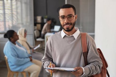 Elinde belgeyle kameraya bakıp vize başvurusu formunu dolduran ve sırasını bekleyen genç, etnik çeşitliliğe sahip bir adam.