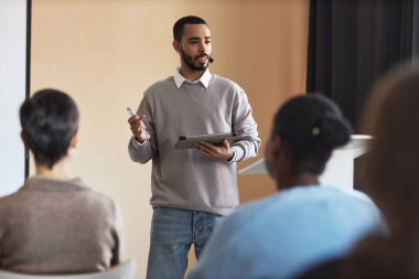 Tablet tabletli genç kendine güvenen koç, beyaz tahtada dururken seminerde veya konferansta izleyicilere sunum noktasını açıklıyor