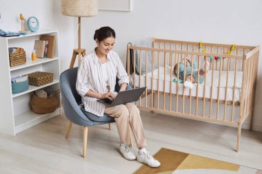 Çalışan genç bir annenin evde dizüstü bilgisayarı kullanırken ve oğlunu izlerken tam boy portresi.