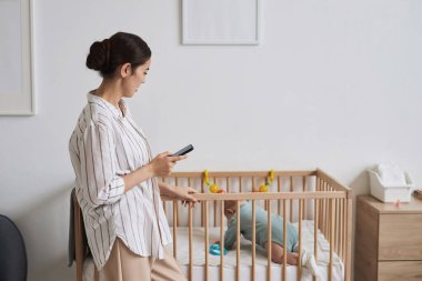 Bebek bakımı ve fotokopi alanı araştırması yaparken elinde akıllı telefon tutan genç annenin yan görünümü.