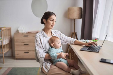 Evden çalışan ve iş yerinde kucağında bebeğini tutan genç bir annenin yan görünüm portresi.