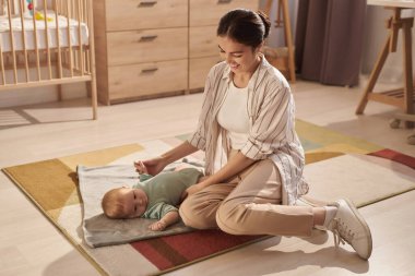 Gülümseyen genç annenin, yumuşak battaniyeli bebekle oynarken yerde oturup, boşluğu kopyaladığı uzun bir portre.
