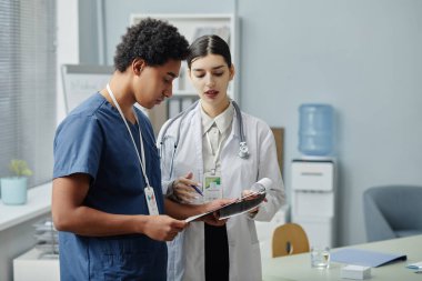 İki genç stajyerin yan görünüş portresi. Ellerinde dosya var ve klinikte hastaların durumunu tartışıyorlar.