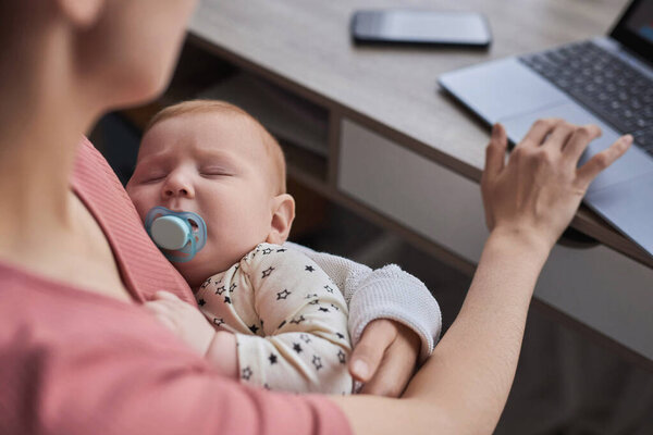 Крупный план работающей матери с помощью ноутбука и с милым спящим ребенком в руках, копировальное пространство
