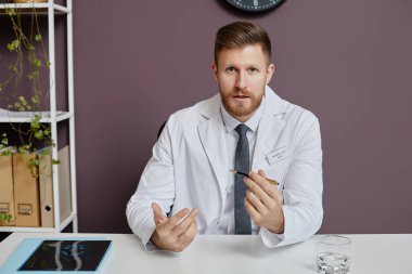 Profesyonel sakallı doktorun portresi klinikteki işyerinde oturup kamerayla konuşuyor, fotokopi çekiyor.