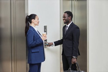 Kafkasyalı bir kadının, kahve fincanı ve takım elbiseli, gülümseyen siyah bir adamın ofis salonundaki asansörde birbirleriyle sohbet ederken orta uzunlukta bir pozu.