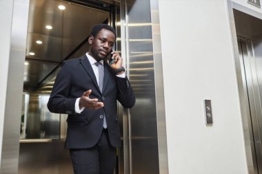 Afrika kökenli Amerikalı iş adamının zarif bir takım elbiseyle iş merkezindeki asansörden çıkarken telefonda konuştuğu düşük açılı bir kare.