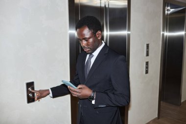 Ofis lobisinde asansör düğmesine basarken iş kıyafeti giymiş siyahi bir adamın telefona baktığı belden yukarı pozlar.
