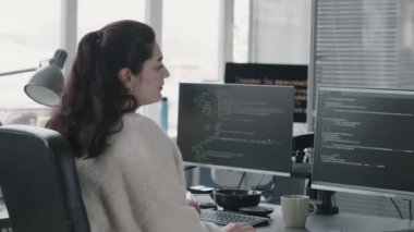 Orta Doğu 'lu genç kadın bilişim uzmanının orta boy portresi kamerada ofis koltuğuna dönüyor ve gülümsüyor, ekranda programlama kodları olan iki masaüstü bilgisayarının önünde oturuyor.