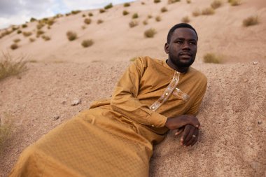 Portrait of african American man wearing long kaftan dress lying on sand dunes in desert and looking away wistful clipart