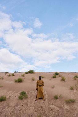 Siyah adamın çölde kum tepecikleri üzerinde dikey açıyla durduğu ve geleneksel kaftan giydiği bir kare.