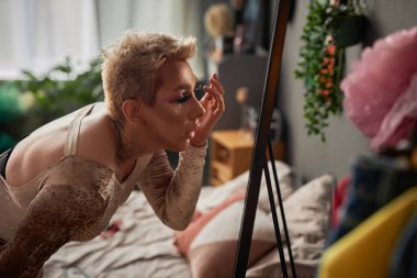 Makyaj yapan, aynaya bakıp takma kirpikleri düzelten şımarık bir gencin yan görünüm portresi.