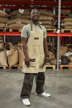 Siyahi bir gencin dikey portresi. Kameraya gülümsüyor ve kahve dükkanında dikilirken önlük giyiyor.