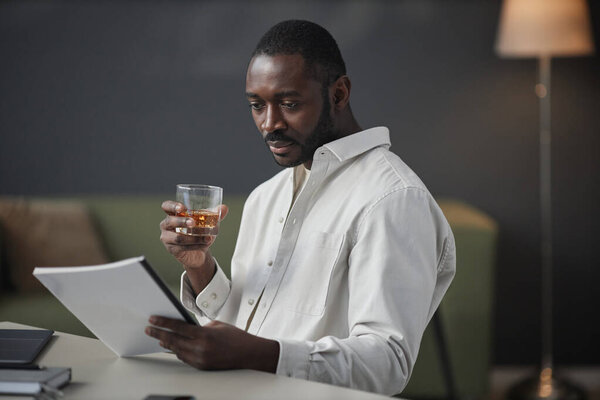 Портрет успешного афроамериканского предпринимателя, читающего документы на рабочем месте в офисе и держащего стакан виски, копировальное пространство