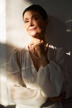 Güneş ışığında beyaz elbise giyen ve kameraya bakan zarif yaşlı gelinin dikey portresi.