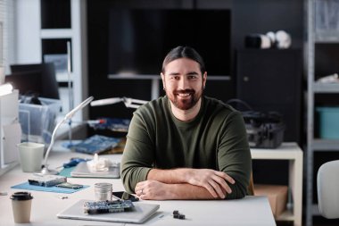 Kameraya bakan sakallı adamın portresi. Teknoloji tamir atölyesinde oturuyor.