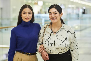 Alışveriş merkezinde alışveriş yapan ve kameraya gülümseyen iki Orta Doğulu kadının belden yukarı portresi.