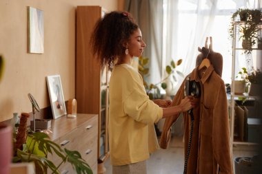 Saçı kıvırcık, kadife ceketli, rahat bir evde giysileriyle ilgilenen genç bir kadının yan görünümü.