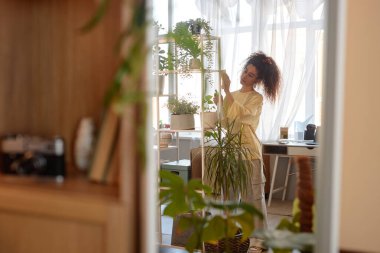 Konforlu ev fotokopi alanındaki canlı yeşil bitkilerle rafları temizleyen genç kadının ayna yansıması.