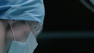 Ameliyat odasında koruyucu gözlük ve yüz maskesi takan konsantre beyaz erkek doktorun orta boy yakın çekimi.